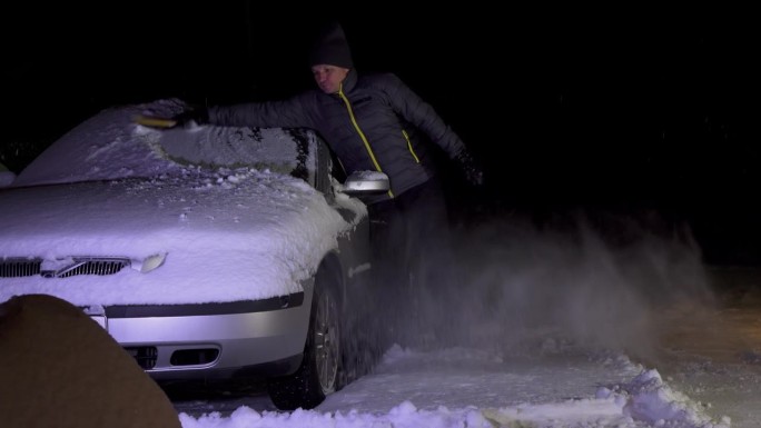 傍晚下班后，一名男子站在车道上清理车上的积雪。一个男人用长长的大刷子打扫雪