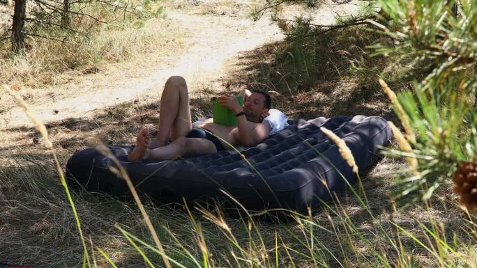 在一个阳光明媚的夏日，一个男人躺在树林里的充气床垫上看书。