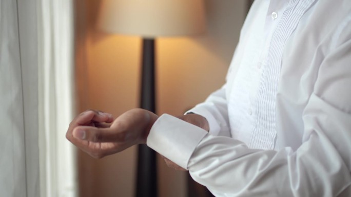 优雅的男人在房间里穿衣服。新郎在准备婚礼时正扣着白衬衫的扣子。
