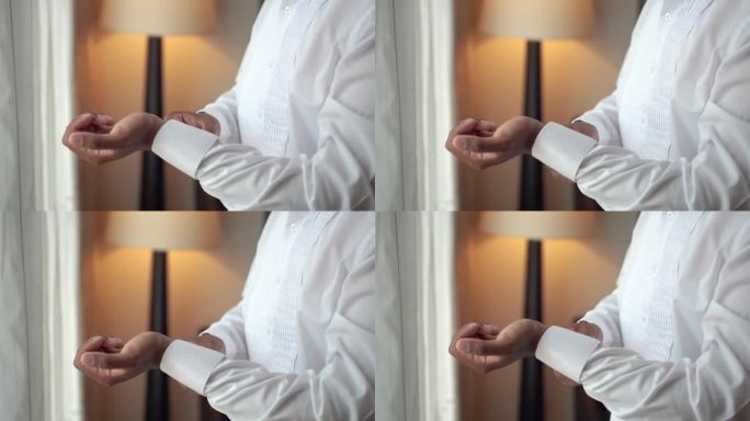 优雅的男人在房间里穿衣服。新郎在准备婚礼时正扣着白衬衫的扣子。