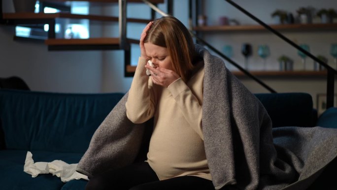 生病的年轻孕妇咳嗽，流鼻涕，坐在家里的客厅里头痛。生病的高加索人的肖像在室内表现出疾病症状。