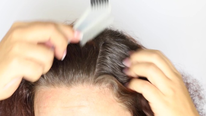 特写:一个无法辨认的女人用鼠尾梳把深棕色的卷发分开，露出头发的根，灰白的头发。