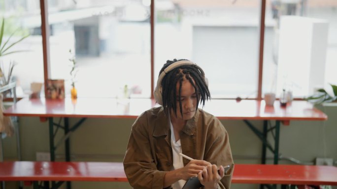 年轻的亚洲男子在咖啡馆工作，拿着平板电脑和文件，一边喝着咖啡，戴着耳机听着音乐。
