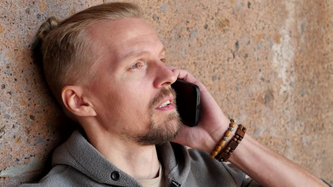 一个英俊的年轻时髦的棕色眼睛和山羊胡子的男人靠在棕色的石墙上用手机说话。一个用智能手机打电话的男人手