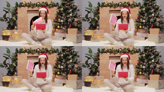 圣诞节的庆祝活动。惊讶的年轻女子穿着温暖的毛衣，戴着圣诞老人的帽子，坐在圣诞树旁的地板上，带着惊讶的