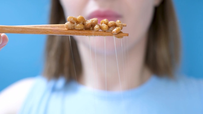 年轻女子用木筷子吃日本纳豆(正面特写，蓝色背景)。