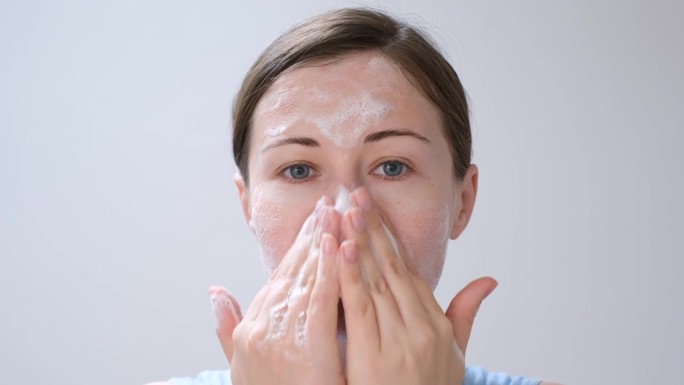 一名年轻女子将洁面泡沫涂抹在脸上(特写，白色背景)。