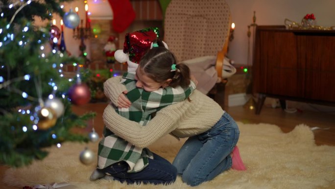 兄妹俩在地毯上的圣诞树旁相对而坐，欢快地拥抱在一起。