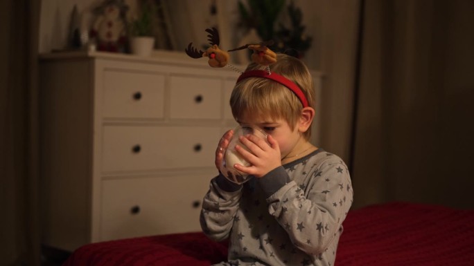 圣诞快乐。有趣的孩子男孩喝牛奶，玩得很开心。新年快乐。兴奋的小孩穿着睡衣坐在床上做鬼脸。戴着红色狂欢