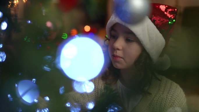 特写镜头。一个戴着圣诞帽的女孩在圣诞树上精心挑选圣诞球。