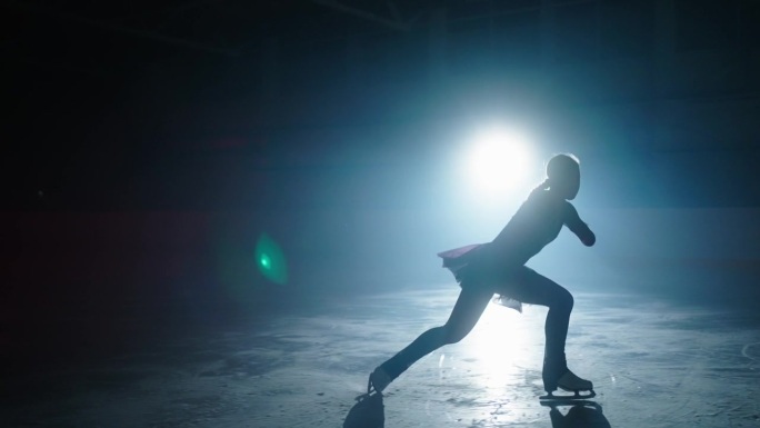 美丽的年轻女子花样滑冰运动员跳舞在溜冰场在黑暗中，慢动作镜头，剪影