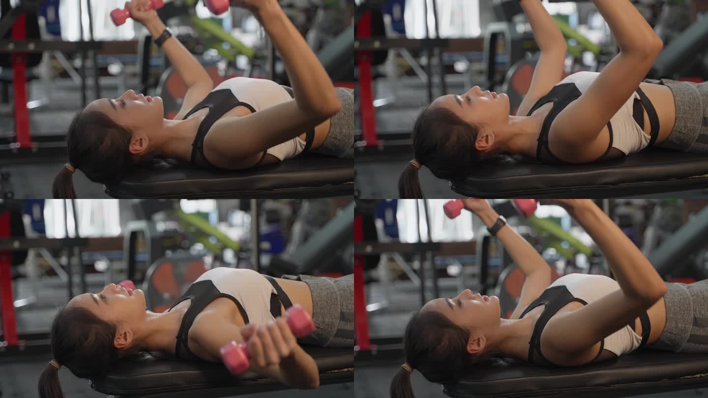 迷人的亚洲女人躺在健身房的长凳上举起小哑铃来暖胳膊