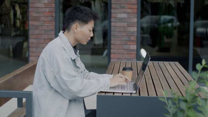 年轻的亚洲男性自由职业者坐在现代街头咖啡馆的桌子上用笔记本电脑工作