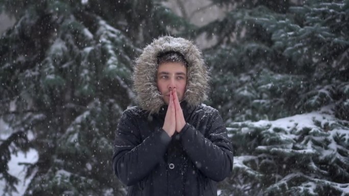 大雪下，一个年轻人站在冷杉树的背景下，用手搓着御寒。一名身穿羽绒服、戴着兜帽的男子站在那里看着镜头。
