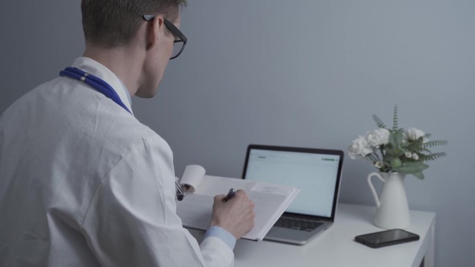 年轻的男医生戴着眼镜，穿着白大褂，坐在办公桌前操作笔记本电脑。医生检查病人的化验结果并将数据输入电脑