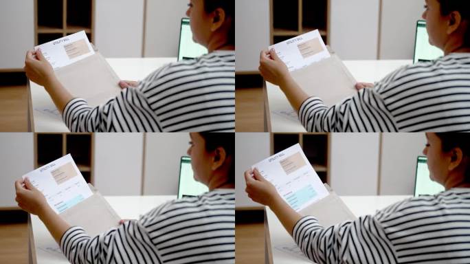 一名妇女在看信封里的水电费账单，上面写着在工作场所通过笔记本电脑在线支付的金额