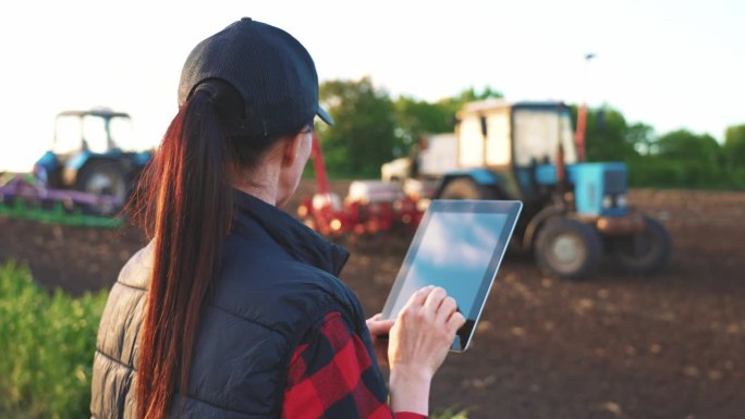 妇女在田间工作拖拉机的背景下，在平板电脑上使用专门的应用程序。农田遥控机械设备。小企业工作理念优化。