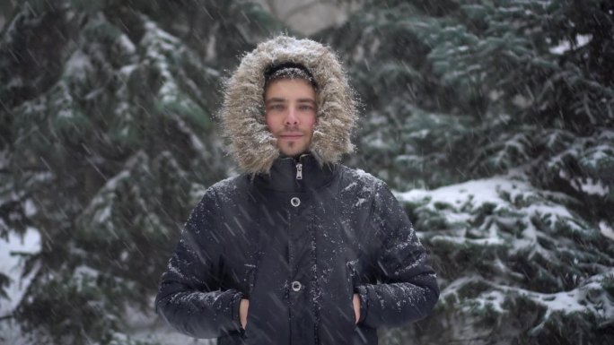 冬天下大雪，一个年轻人站在冷杉树的背景下。一名身穿羽绒服、戴着兜帽的男子站在那里看着镜头。4 k