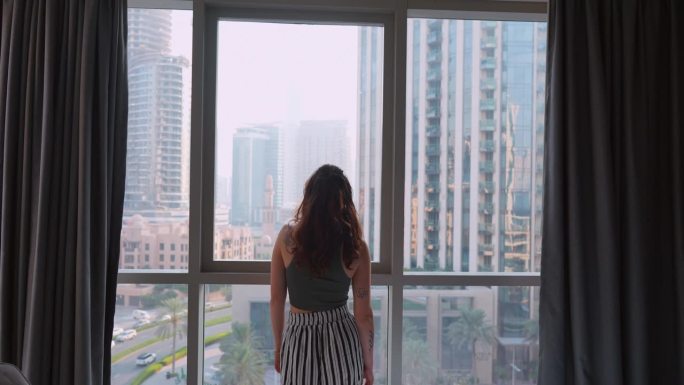 一名女子站在窗边，从她的公寓里看着迪拜的城市景观