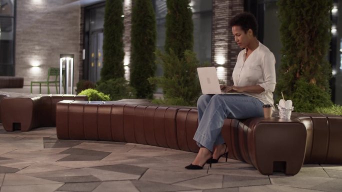 黑人女商人晚上坐在长凳上用笔记本电脑写报告