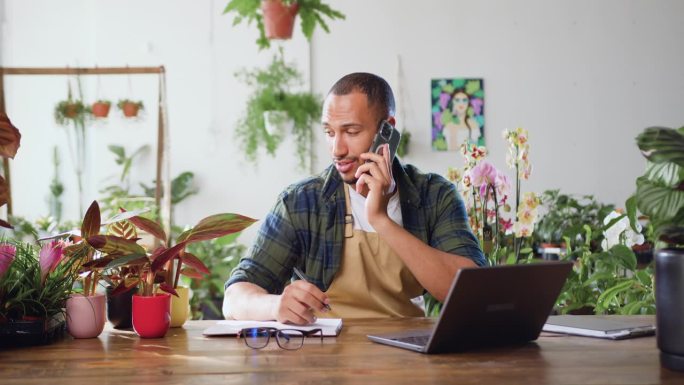 温室植物学家，男性植物工作者，坐在自己的花店里，在笔记本电脑上打字，和客户使用电话。花店的一位花商从
