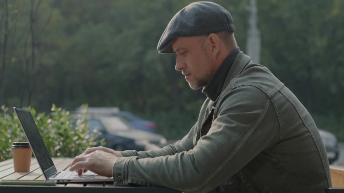 在城市背景下，坐在露天咖啡馆里用笔记本电脑打字的积极的商人的侧视图