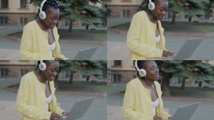 快乐的自由职业者非洲裔美国女孩使用笔记本电脑，戴着耳机听音乐，享受户外街头的歌曲