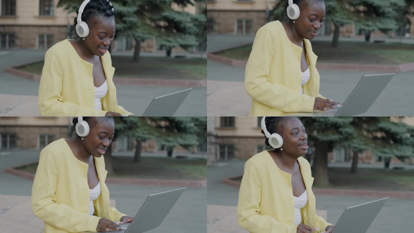 快乐的自由职业者非洲裔美国女孩使用笔记本电脑，戴着耳机听音乐，享受户外街头的歌曲