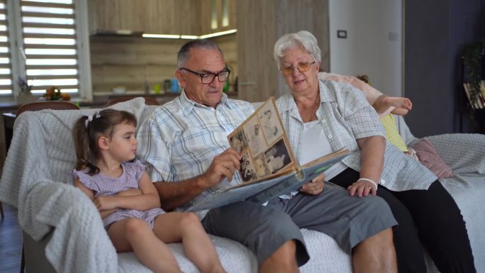 上了年纪的祖父母和大孙女坐在沙发上看相册，双胞胎小女儿在后面玩耍