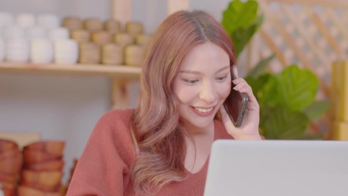 美丽的商业企业家亚洲年轻女子在网上卖花瓶花与笔记本电脑接电话，得到好消息，在家里庆祝她的工作成功愉快