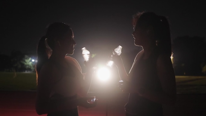 女人晚上户外健身喝水灯光逆影两个女人