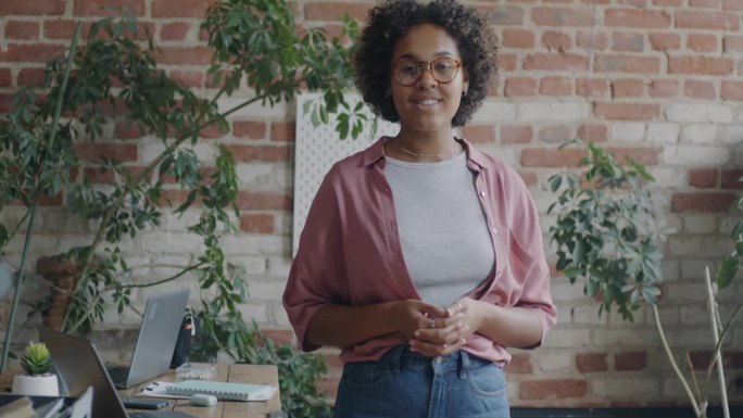 快乐的非裔美国女企业家转向镜头，微笑着站在创意loft风格的办公室里