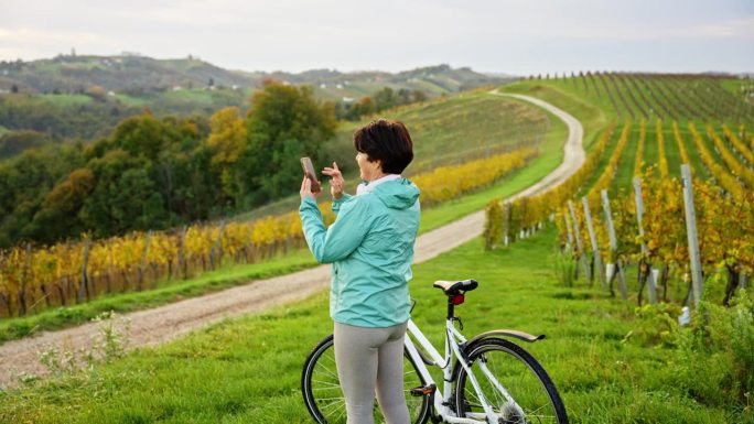 快乐的成熟女子站在自行车旁，用智能手机在乡村拍摄葡萄园