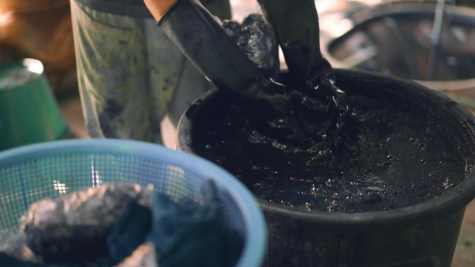 当地人戴着黑色橡胶手套，在装有颜料溶液的罐子里用深蓝色颜料染布，并用手挤压布料，使染色过程均匀，这是