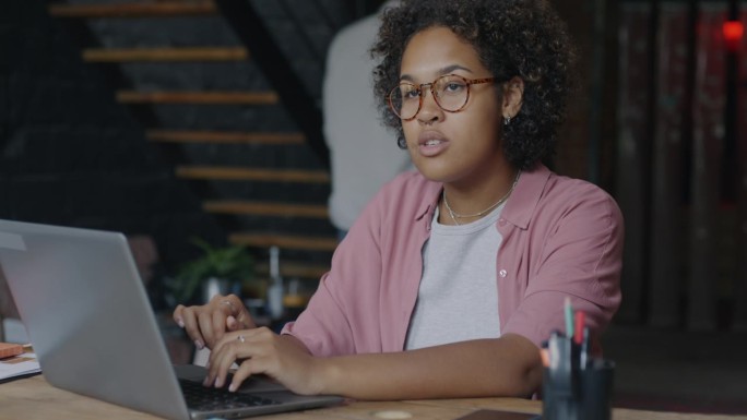 快乐的非裔美国女性办公室职员的肖像，在创造性的开放空间工作场所用笔记本电脑打字