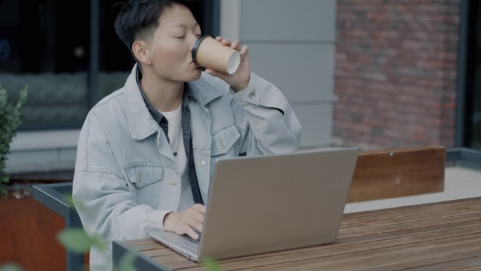 亚洲学生在网上学习，坐在街边的咖啡馆里，用笔记本电脑学习，喝着咖啡