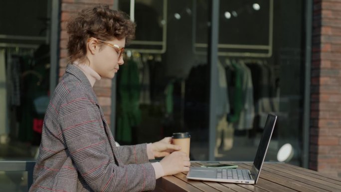 侧视图，自信的年轻女子使用笔记本电脑打字，喝咖啡，午休时间在街边咖啡馆工作