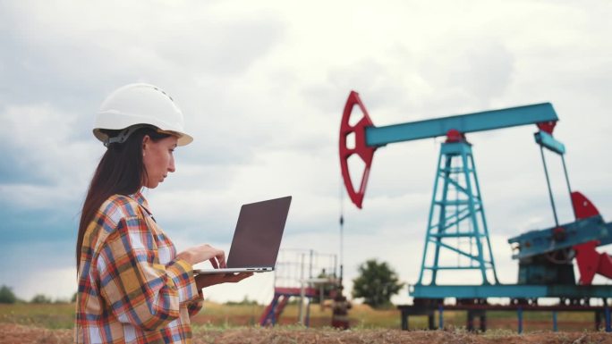 石油工业。工程师站在油田钻井站旁边，用笔记本电脑在键盘上输入文本。石油生产企业财务概念。天然气生产。