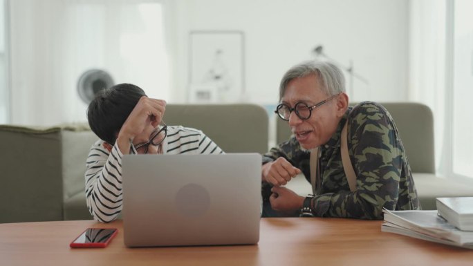 数字时代的家庭支持:亚洲男性自由职业者在舒适的家庭环境中与年长的父亲交谈和工作。
