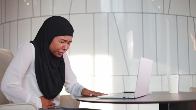 沮丧的多元文化女性戴着黑色头巾，在工作场所胃部不适时用手触摸腹部。上班族营养不规律的后果。办公室工作