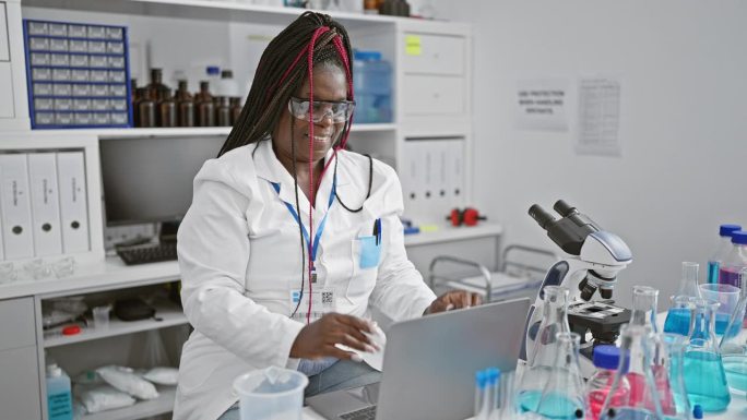 激动的非裔美国女科学家，摇着她的辫子和眼镜，在实验室里用笔记本电脑探索医学奇迹时，打出了胜利庆祝的手