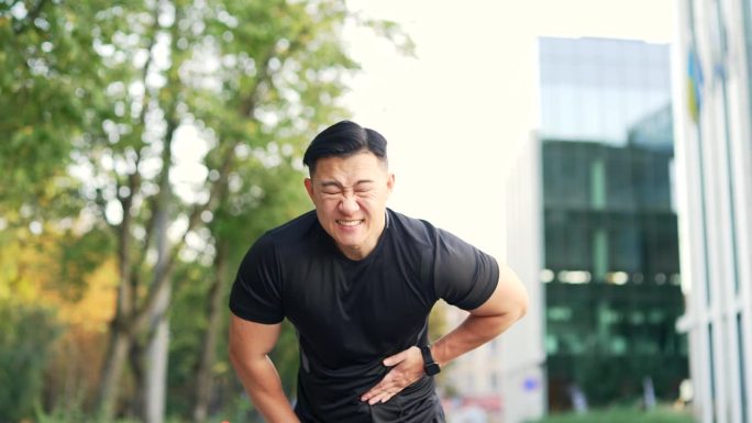 一名亚洲运动员在城市街道上跑步时感到胃痛。穿黑色t恤的男运动员有胃灼热