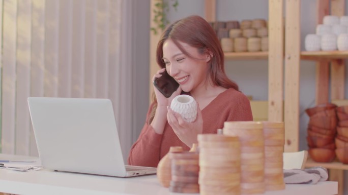 美丽的商业企业家亚洲年轻女子在网上卖花瓶花与笔记本电脑接电话，得到好消息，在家里庆祝她的工作成功愉快