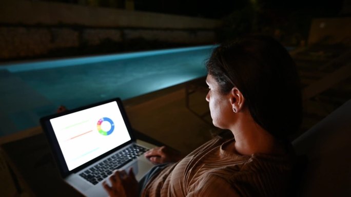 晚上在游泳池边用笔记本电脑工作的女人。女性通过网络平台进行理财