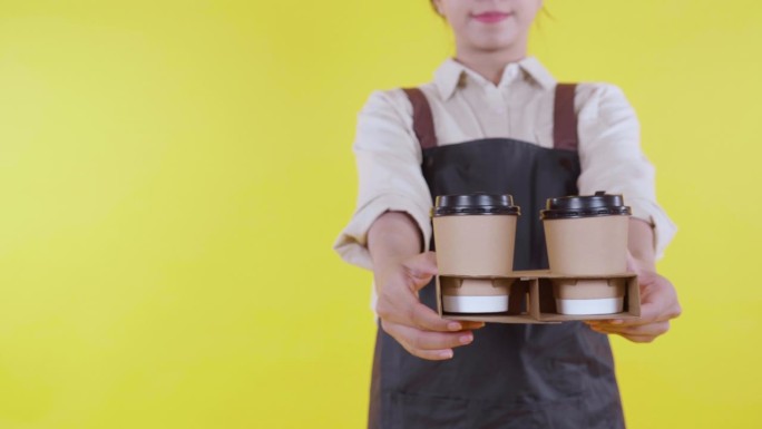 年轻的亚洲咖啡师的手，穿着围裙，拿着黄色背景的咖啡杯，服务员或企业家兴奋地拿着咖啡，小生意或创业，咖