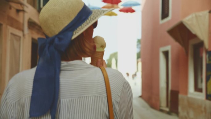 在克罗地亚村庄的街道上行走时，女游客吃着新鲜的甜筒冰激凌的后视图