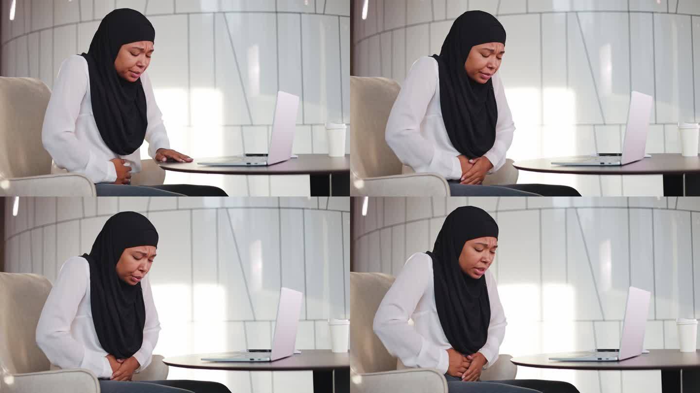 在现代办公室使用手提电脑工作时感到胃部不适的不快乐的成熟女性。戴着黑色头巾的多种族女经理因食物中毒而