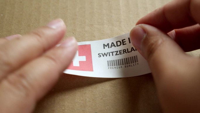 用手将“瑞士制造”标志标签贴在带有产品优质条形码的运输箱上。制造和交付。产品出厂进出口。