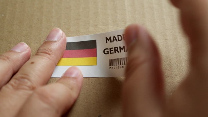 用手将德国制造的旗帜标签贴在带有产品优质条形码的运输箱上。制造和交付。产品出厂进出口。
