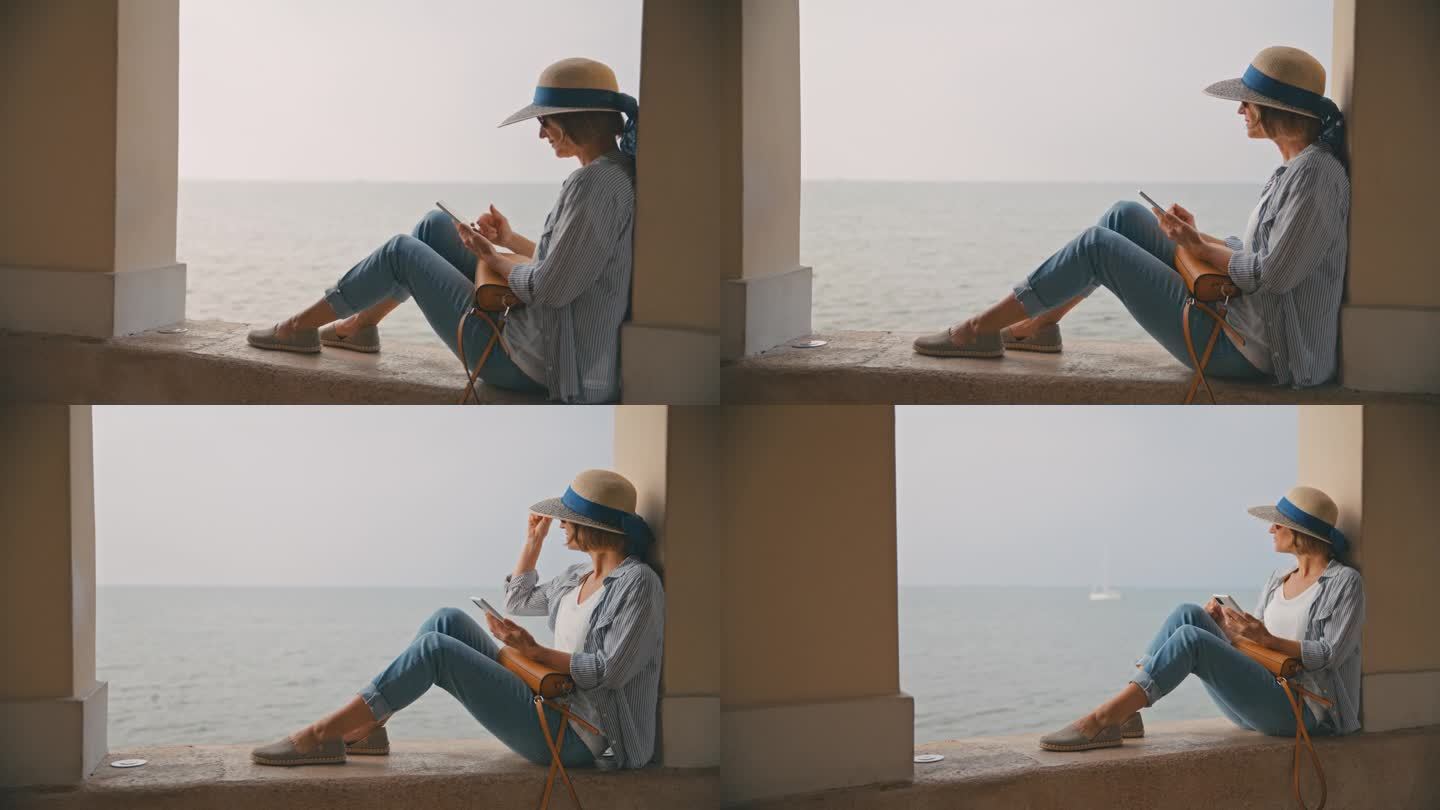 悠闲的女游客坐在克罗地亚柱廊的挡土墙上，一边用智能手机一边看海景